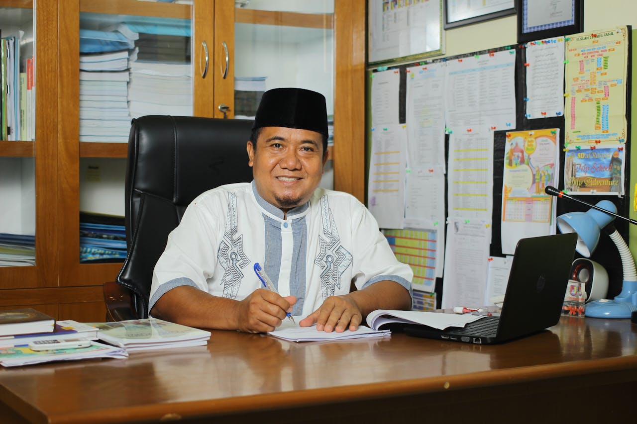 An Indonesian teacher in traditional attire, smiling at his desk with documents in an office setting.